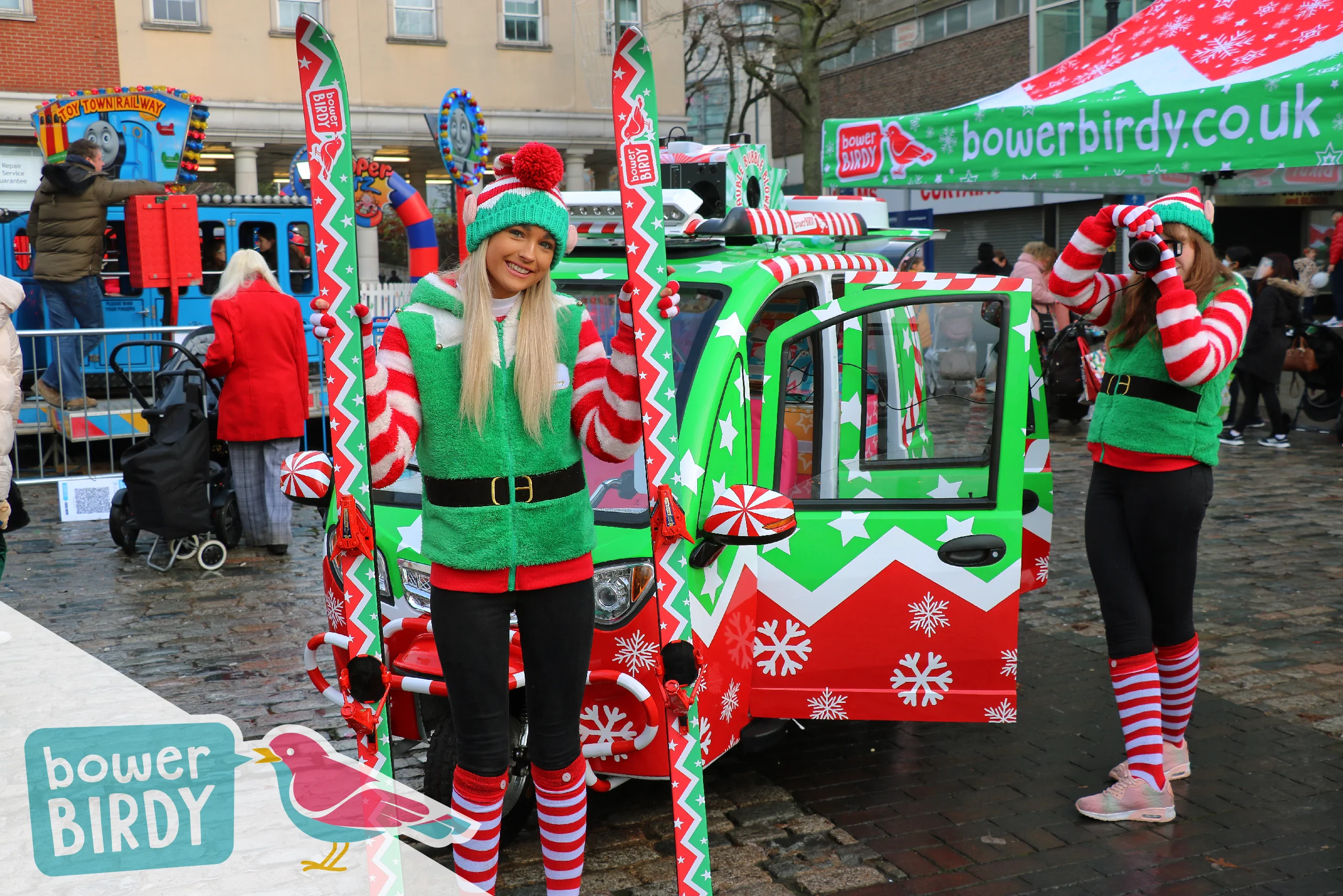Elf Car Christmas Event : A Bowerbirdy elf poses with a pair of red and green elf skis in from of the Elf Car, while another elf takes a photo.