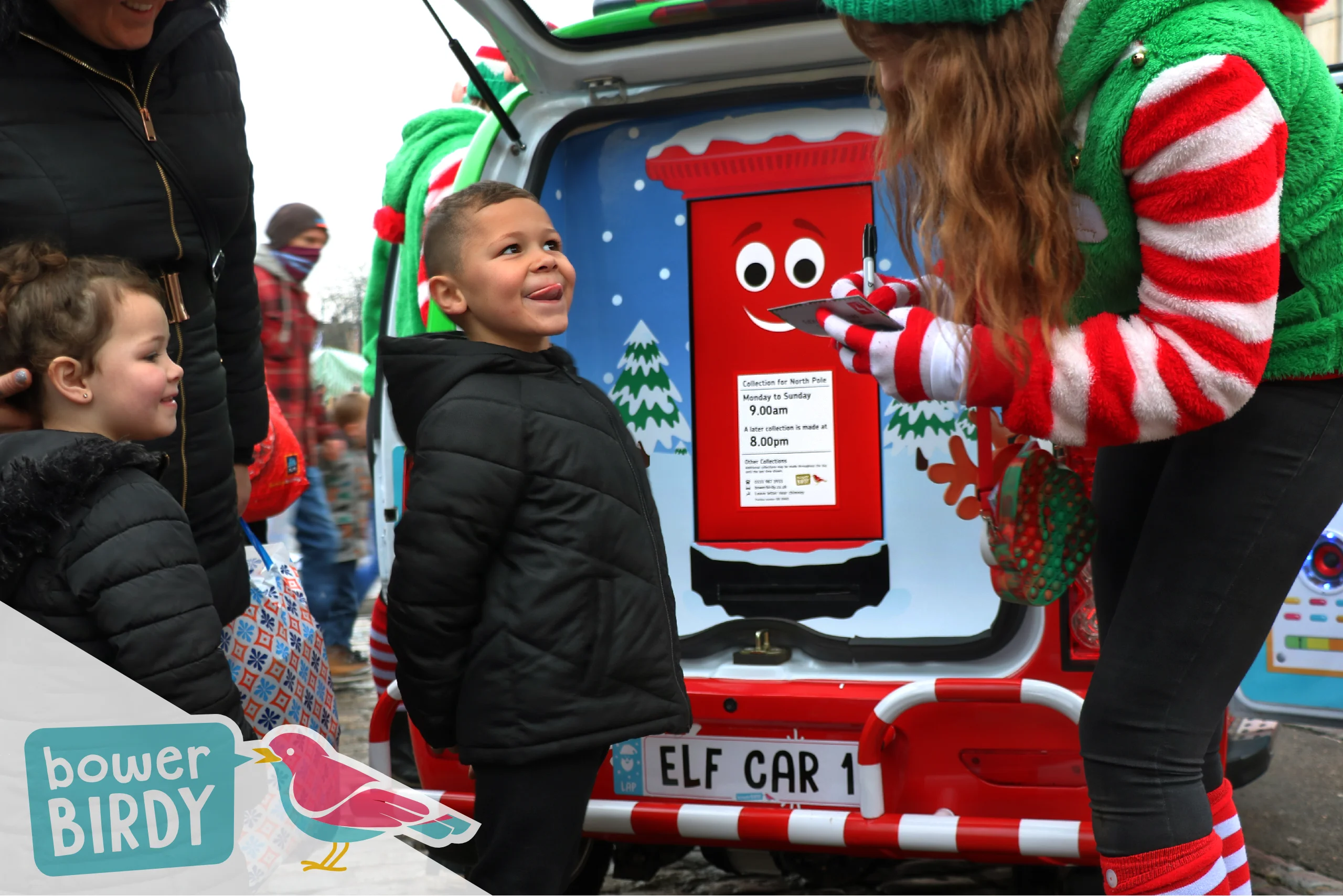 Elf Car Christmas Event: A boy pulls a cheeky face while a Bowerbirdy Elf helps to write his christmas list, in front of the Enchanted Postbox