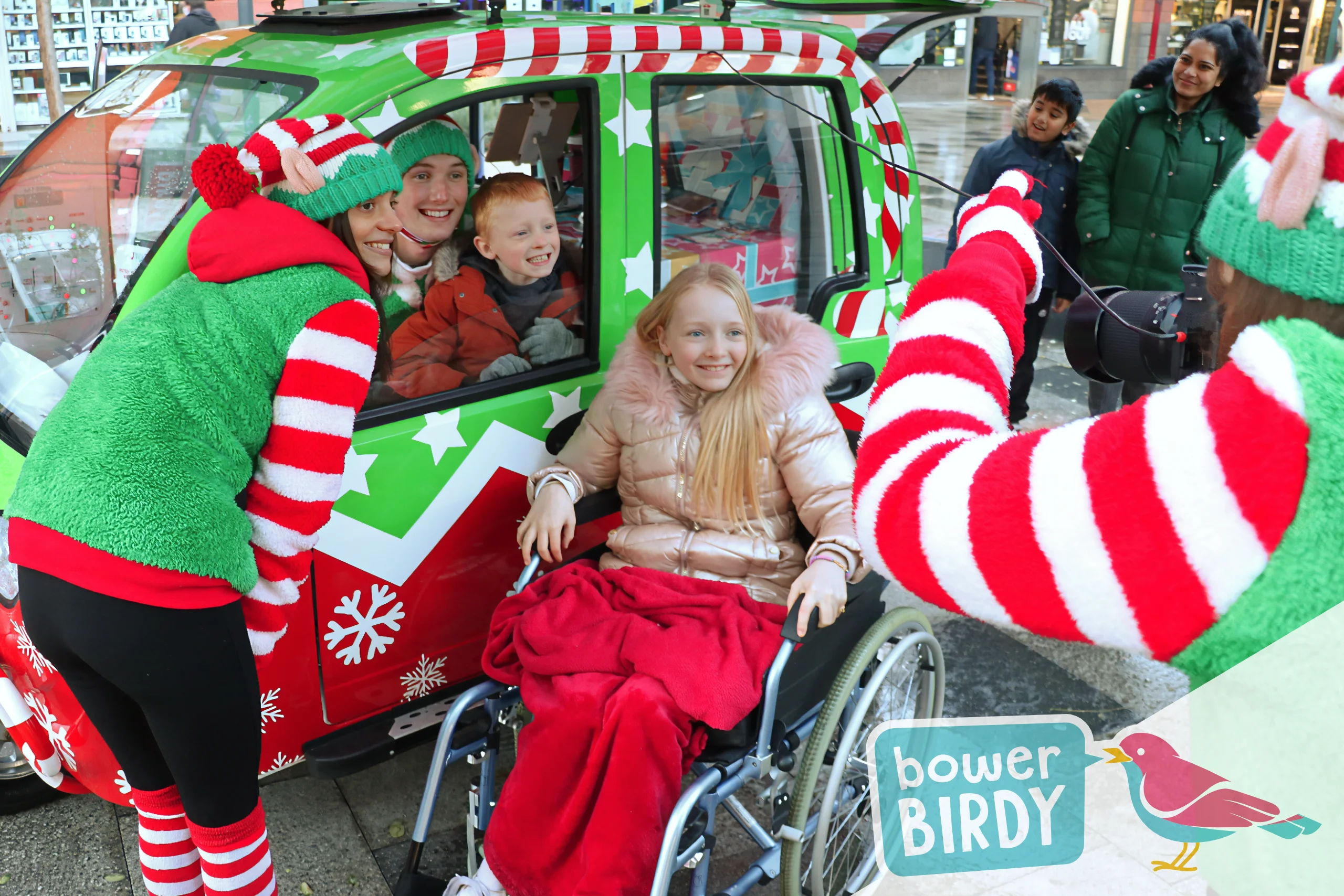 Elf Car Christmas Magic in Stevenage: A boy and a Bowerbirdy elf inside the Elf Car, and a Bowerbirdy Elf and a girl in a wheelchair outside of the Elf Car all smile for a photo, being taken by a third elf.