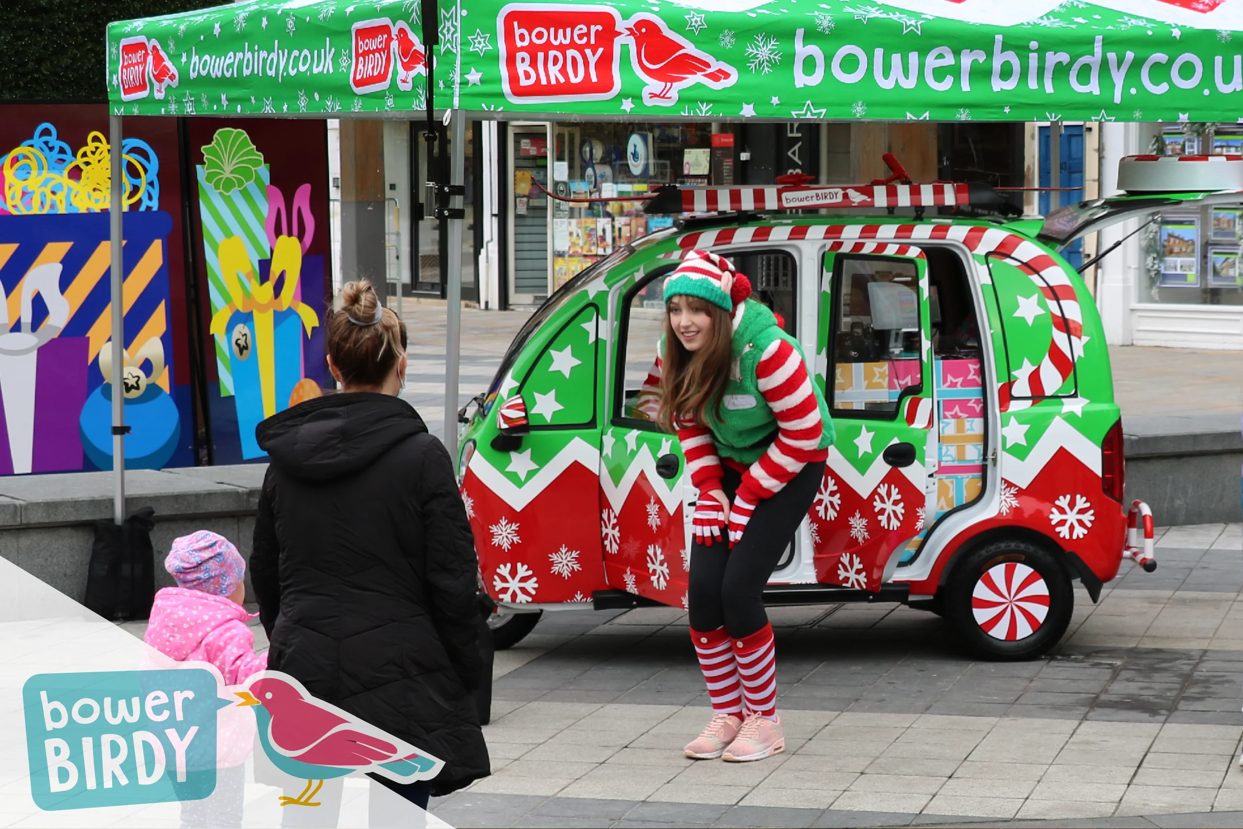 A Bowerbirdy elf talks to a young girl and her mum in front of the Elf Car